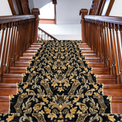 View looking up a straight wooden stairway covered in an ornate black and gold stair runner. 