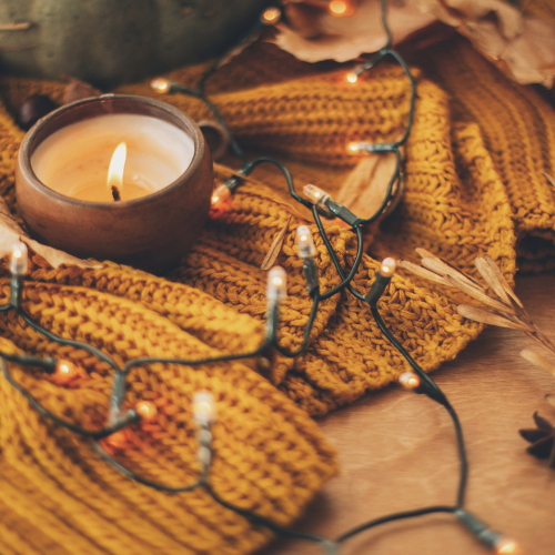 Candle in a fall setting with string lights and a mustard-colored knit blanket for cozy fall ambiance.